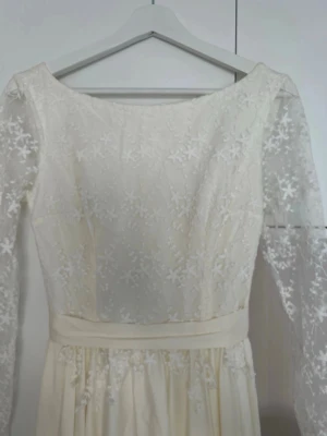 Brudklänning med spetsdetaljer - Fin cremevit brudklänning med transparenta spetsärmar och blommiga broderier på överdelen. Klänningen har en markerad midja med ett band, djup urringning i ryggen och ett flytande långt kjolparti. Mycket fint skick men kan behöva en kemtvätt. Inköpt för 14 999kr.