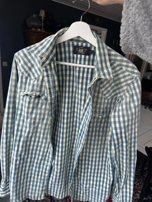 Rutig skjorta från Ralph Lauren - Säljer en klassisk rutig skjorta från Ralph Lauren i blått och vitt. Skjortan har lång ärm, två bröstfickor med knappar och en avslappnad passform. Perfekt för dig som gillar tidlös stil med en modern twist.