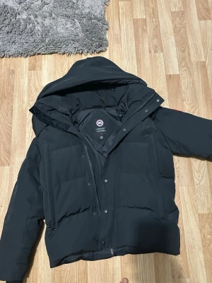 Svart pufferjacka från Canada Goose - Säljer en svart pufferjacka från Canada Goose i storlek M. Jackan har huva, dragkedja och knappar framtill samt flera praktiska fickor. Perfekt för kalla dagar och riktigt snygg till streetwear-looken.