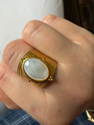 Guldfärgad ring med pärlemor justerbar - Säljer en bred guldfärgad ring med en oval pärlemorsten i mitten. Ringen har detaljerade mönster runt stenen och en lyxig vintagekänsla. Perfekt för dig som gillar statement-smycken och unika accessoarer. Justerbar, stainless