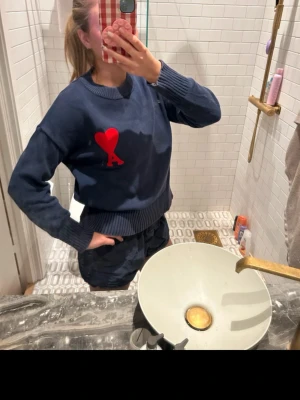 Ami paris tröja - Cool mörkblå sweatshirt från AMI Paris med stor röd hjärtlogo och bokstaven A på bröstet. Tröjan har ribbade muddar vid ärmslut och nederkant samt rund halsringning. Perfekt för dig som gillar streetstyle och snygga detaljer.