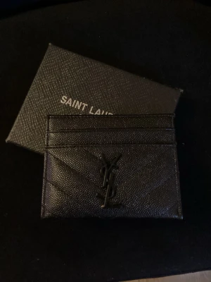 Svart kortfodral från Saint Laurent/ YSL wallet/ Saint laurent wallet/ Wallet - Snyggt svart kortfodral från Saint Laurent i präglat läder med ikoniska YSL-loggan framtill. Fodralet har flera kortfack och stilrena sömmar i chevronmönster. Perfekt för dig som vill ha en lyxig och stilren accessoar.