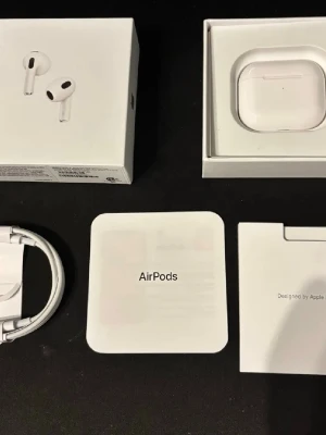 Apple AirPods med laddningsetui - Säljer ett par vita Apple AirPods med trådlöst laddningsetui. Hörlurarna har en stilren och minimalistisk design i blank plast och levereras med originalförpackning, laddningskabel och manualer. Perfekta för musik, samtal och smidig anslutning till Apple-enheter.