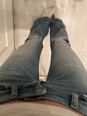 Ljusblå slitna bootcut jeans - Säljer ett par ljusblå bootcut jeans med slitningar och hål vid knäna. Jeansen har klassisk femficksdesign och stängs med knapp och dragkedja. Perfekta för en avslappnad och trendig look.