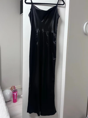 Svart långklänning i satin - Snygg svart långklänning med tunna axelband och slät, glansig satinlook. Klänningen har en enkel och elegant siluett med lätt v-ringad halsringning. Perfekt för dig som vill ha en stilren och trendig look.