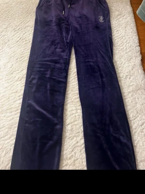 Mörkblå velourbyxor från Juicy Couture - Säljer ett par mörkblå mjukisbyxor i velour från Juicy Couture. Byxorna har raka ben, dragsko i midjan och fickor. På ena benet finns Juicy Couture-loggan och baktill står det 'Juicy' i glittriga stenar. Perfekta för en chill och trendig look.