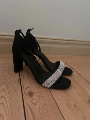 Svarta Glittriga Klackskor✨ - Festliga svarta klackskor från XIT med bred rem framtill täckt av glittrande strass. Skorna har en chunky klack och justerbar rem runt ankeln. Perfekta för att lyfta vilken outfit som helst med lite extra bling. Hade dessa skor på min bal men har tyvärr inte fått användning för dom utöver det. Super bekväma för att vara klackskor. ✨💕
