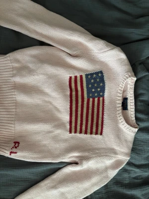 Vit stickad tröja Ralph Lauren - ❗️INTRESSEKOLL ❗️Vit stickad tröja från Ralph Lauren med amerikansk flagga i blått, rött och vitt på bröstet. RL-broderi i rött vid nederkanten. Klassisk rund halsringning och ribbstickade muddar vid ärmslut och midja. Perfekt för dig som gillar preppy stil. 