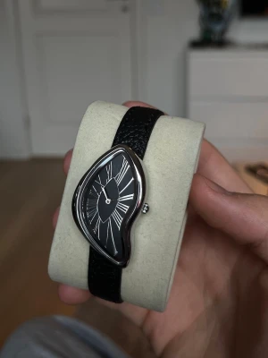 Svart klocka - Unik klocka med svart läderband. Urtavlan verkar smelt och är svart med vita romerska siffror och tunna visare. Perfekt för dig som gillar konstnärliga och annorlunda accessoarer.