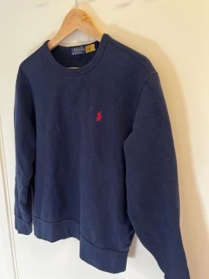 Ralph Lauren Crewneck Sweatshirt - Kvalitétströja från Polo Ralph Lauren säljes i fint skick  Säljer en klassisk marinblå sweatshirt från Polo Ralph Lauren i storlek M. Tidlös modell med rund hals och röd broderad logotyp på bröstet.  Tröjan är i fint begagnat skick utan hål eller fläckar. Normal passform och mjuk insida som gör den skön att bära. Perfekt till både vardag och mer uppklätt casual helg.  Köpt på CareofCarl 2025 för 2200kr. Använd endast ett fåtal gånger. 