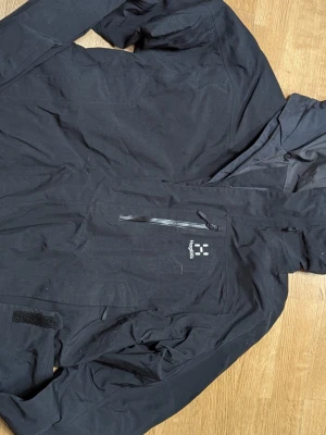 Svart vindjacka från Haglöfs - Snygg svart vindjacka från Haglöfs med huva och dragkedja på bröstet. Jackan har minimalistisk design, diskret logga på bröstet och justerbara ärmslut med kardborre. Perfekt för blåsiga dagar och outdoor-äventyr.