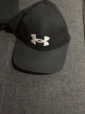 Svart keps från Under Armour - Snygg svart keps från Under Armour med böjd skärm och stort vitt logotryck framtill. Klassisk sexpanel-design och justerbar passform. Perfekt accessoar för en sportig och avslappnad stil.