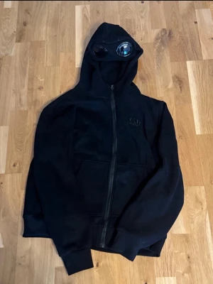 Svart hoodie med goggles från C.P. Company - Svart hoodie från C.P. Company med hel dragkedja och ikoniska goggles i huvan. Broderad logga på bröstet och känguruficka framtill. Perfekt för dig som gillar streetwear och unika detaljer. Materialet känns mjukt och bekvämt. Dock är den lite oversized så de fungerar på storlek L! Ni kan gärna komma med bud!