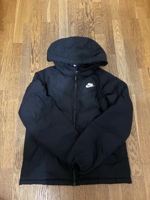 Svart pufferjacka från Nike - Säljer en svart pufferjacka från Nike med huva och dragkedja framtill. Jackan har en klassisk Nike-logga på bröstet och är tillverkad i ett quiltat material som håller dig varm. Perfekt för kalla vinterdagar och enkel att matcha med olika outfits.