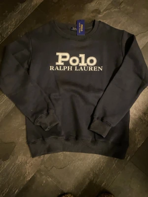 Marinblå  sweatshirt från Polo Ralph Lauren M - Säljer en marinblå sweatshirt från Polo Ralph Lauren med stor vit logga på bröstet. Tröjan har rund halsringning, ribbade muddar vid ärmslut och nederkant samt en klassisk, avslappnad passform. Perfekt för en stilren och enkel look.