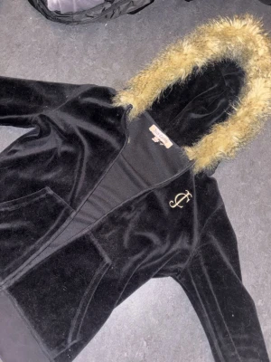 Juicy Couture svart velour hoodie - Juicy Couture hoodie i svart velour med stor luva kantad av fuskpäls. Klassisk JC-logga broderad på bröstet och två framfickor. Perfekt för dig som gillar en lyxig och avslappnad stil.