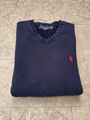 Mörkblå stickad tröja Polo Ralph Lauren - Klassisk mörkblå stickad tröja från Polo Ralph Lauren med rund halsringning och ribbade muddar. Tröjan har den ikoniska röda logotypen broderad på bröstet. Tillverkad i mjukt material som ger en skön och avslappnad passform.