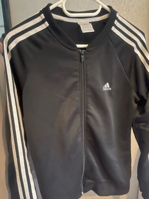 Svart träningsjacka från Adidas - Snygg svart träningsjacka från Adidas med klassiska vita ränder längs ärmarna och dragkedja framtill. Adidas-logga på bröstet och glittrig text på ryggen. Perfekt för sportiga tillfällen.