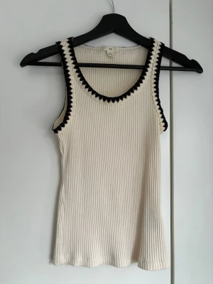 Ribbad linne från H&M med kontrastkant - Säljer ett ribbat linne från H&M i offwhite med svarta virkade kontrastkanter runt halsringning och ärmhål. Linnet har en rund hals och är figurnära i modellen. 