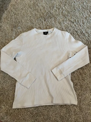 Vit långärmad tröja från H&M - Säljer en enkel och stilren vit långärmad tröja från H&M. Tröjan har rund halsringning och är tillverkad i ett ribbat, mjukt material som känns skönt mot huden. Perfekt basplagg att matcha med olika outfits.