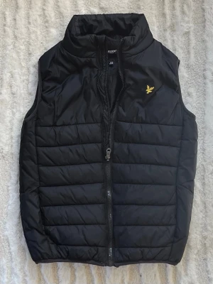 Svart dunväst från Lyle & Scott, strl 9/10y - Svart dunväst från Lyle & Scott med gul logotyp på bröstet. Bra skick. Strl ungdom 9/10 år. Längd axel-nederkant ca 50cm.