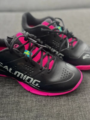 Salming svarta inomhusskor rosa detaljer - Helt nya oanvända  inomhusskor från Salming i svart med rosa detaljer och sula i gummi. Skorna har ventilerande meshpartier, svart snörning och Salming-logga på sidan. Perfekta för innebandy eller andra inomhussporter där grepp och stabilitet behövs.