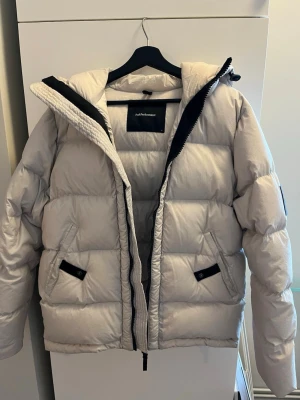 Ljus dunjacka från Peak Performance - Säljer en ljus dunjacka från Peak Performance med svart detaljer och dragkedja. Jackan har två fickor med svarta knappar och en hög krage. Perfekt för kalla vinterdagar och har en modern, puffad design.