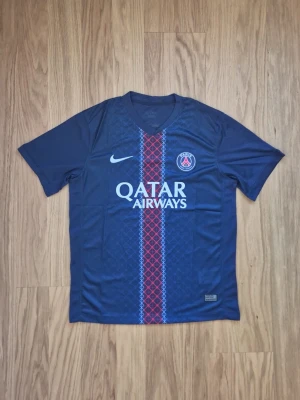  2026 PSG hemma matchtröja blå Nike - Säljer en blå Paris Saint-Germain matchtröja från Nike med Qatar Airways som sponsor. Tröjan har korta ärmar och ett snyggt mönster i rött och vitt längs mitten. PSG-loggan och Nike-loggan är broderade på bröstet. Perfekt för fotbollsfans!