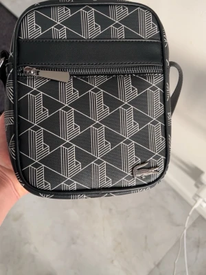 Svart axelväska från Lacoste - Säljer en svart axelväska från Lacoste med vitt geometriskt mönster och silverfärgad dragkedja. Väskan har en justerbar axelrem och ett litet Lacoste-emblem i metall på framsidan. Materialet är tålig syntet och designen är stilren och modern.
