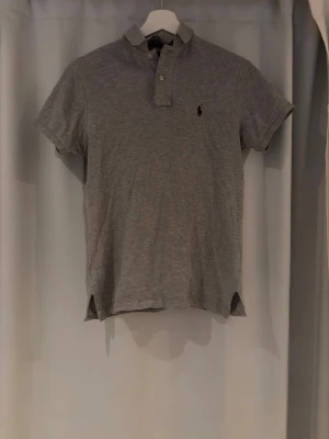 Grå piké från Ralph Lauren - Klassisk grå pikétröja från Ralph Lauren med korta ärmar och liten broderad logga på bröstet. Tröjan har krage och knappar framtill, tillverkad i mjuk bomull för en bekväm känsla. Perfekt till jeans eller shorts för en clean look.