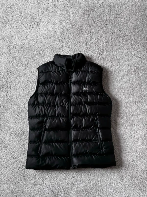 Svart dunväst från Lacoste - Snygg svart dunväst från Lacoste med klassisk ståkrage och dragkedja framtill. Västen har två fickor med dragkedja och den ikoniska gröna krokodilloggan på bröstet. Perfekt för lager på lager och streetwear-vibe.