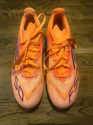 Adidas F50 orange fotbollsskor - Säljer ett par grymma Adidas F50 fotbollsskor i en cool orange färg med lila detaljer och snörning. Skorna har en smal och aerodynamisk form. Materialet är syntetiskt och lätt, vilket ger bra känsla på planen.