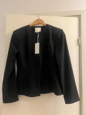 Svart kavaj med knytning från NA-KD - Stilren svart kavaj från NA-KD med dekorativa knytningar framtill och rund halsringning. Kavajen har långa ärmar och en rak passform som ger ett elegant intryck. Materialet är slätt och känns bekvämt mot huden. Perfekt för dig som vill ha en unik twist på klassiska kavajer.
