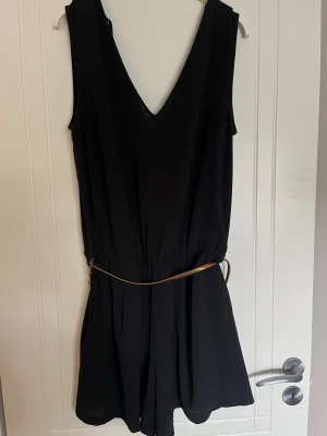 Svart ärmlös jumpsuit med V-ringning - Svart jumpsuit med djup V-ringning både fram och bak, samt knytband i ryggen. Den är ärmlös och har en tunn, guldfärgad metall midjebälte som accentuerar midjan. Materialet ser ut att vara lätt och flowy, perfekt för en cool och avslappnad look.