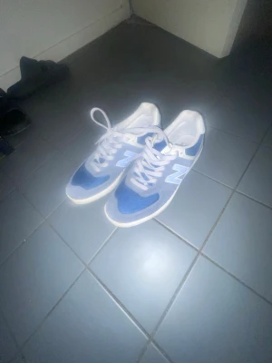 Blå New Balance sneakers med vita detaljer - Snygga blå New Balance sneakers med vita detaljer och klassisk N-logga på sidan. Skorna har vita snören och en låg profil. Ovandelen är i mesh och syntetmaterial som ger en sportig look. Perfekta för dig som vill ha stilrena och bekväma sneakers.