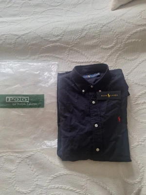 Helt ny  skjorta (aldrig använd) -  Säljer en stilren navyblå Ralph Lauren skjorta med röd logga och vita knappar. Fick den i present men tyvärr passar den inte. • Storlek: M • Skick: Ny (oanvänd) • Nypris: ca 1899 kr • Mitt pris: 899 kr Perfekt till sommaren – funkar både uppklätt och casual. Snabb affär prioriteras, kan skickas direkt eller mötas upp. Hör av dig vid frågor!