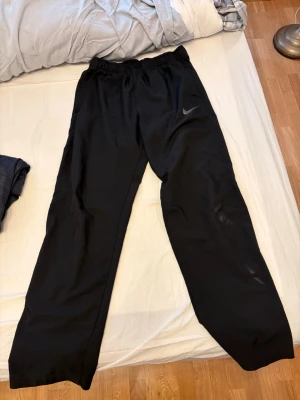 Svarta woven pants från Nike - Säljer ett par svarta woven pants från Nike med elastisk midja och diskret Nike-logga på ena benet. Byxorna har en avslappnad passform och är tillverkade i ett bekvämt material som passar perfekt för chill eller träning.