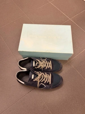 Philippe Model blå sneakers - Snygga blå sneakers från Philippe Model med vita detaljer och gråa skosnören. Skorna har en klassisk låg siluett och är tillverkade i en mix av mocka och textil. Perfekta för dig som gillar stilrena och trendiga sneakers.