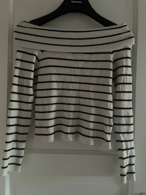 Randig offshoulder topp från H&M - Säljer en ribbad offshoulder topp från H&M i vitt med svarta horisontella ränder. Toppen har lång ärm och en bred, vikt kant över axlarna för en snygg och trendig look. Perfekt för dig som gillar stilrena och moderna plagg.