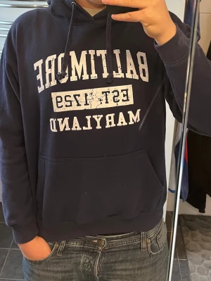 Mörkblå hoodie från ESY Surf Co. - Mörkblå hoodie från ESY Surf Co. med vit text 'BALTIMORE EST. 1729 MARYLAND' på bröstet. Klassisk känguruficka framtill och justerbar huva med snörning. Perfekt för en avslappnad och sportig stil.