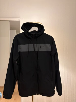Svart vindjacka från Under Armour - Snygg svart vindjacka från Under Armour med huva och dragsko. Jackan har en grå rand över bröstet med logga, hel dragkedja framtill och meshfoder för extra komfort. Perfekt för blåsiga dagar och sportiga outfits.