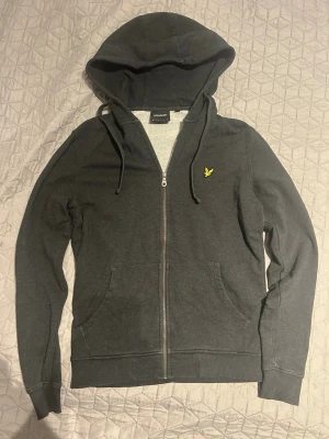 Mörkgrå hoodie från Lyle & Scott - Snygg mörkgrå hoodie från Lyle & Scott med dragkedja framtill, huva med snörning och två fickor. Klassisk gul logga på bröstet. Perfekt för en avslappnad och stilren look.