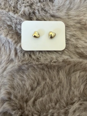 Guldiga hjärtformade örhängen - Säljer ett par söta örhängen i hjärtform med blank guldfinish. Perfekta för dig som gillar minimalistiska och stilrena accessoarer. Örhängena är små och diskreta, vilket gör dem lätta att matcha med olika outfits. Materialet är metall med guldfärgad yta.