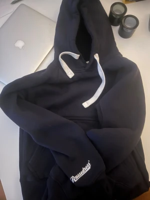 Svart hoodie från Russedress - Svart hoodie från Russedress med vit logga på ärmen och vita snören i huvan. Klassisk passform med stor ficka framtill och ribbade muddar. Perfekt för chill dagar och enkel att matcha med jeans eller joggers. Perfekt för våren 🍃🍃