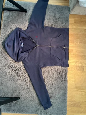 Mörkblå hoodie från Polo Ralph Lauren - Mörkblå hoodie från Polo Ralph Lauren med dragkedja och två stora fickor framtill. Klassisk röd logga broderad på bröstet och mjukt fleecefoder på insidan. Perfekt för chill dagar och snygg att matcha med jeans eller joggers.