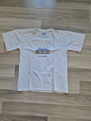 Vit Lanzarote t-shirt med motiv - Vit t-shirt med rund hals och korta ärmar. På bröstet finns ett färgglatt motiv av delfiner och texten 'LANZAROTE'. Materialet ser ut att vara bomull och modellen är klassisk och rak. Perfekt för dig som gillar resor och coola prints.