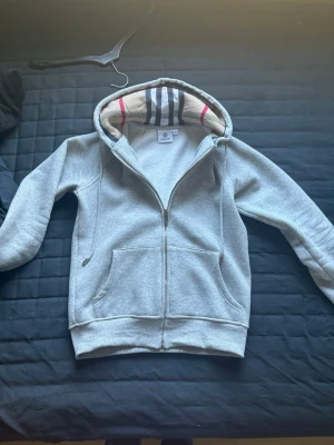 Grå zip hoodie med randig huva - Snygg grå zip hoodie från Burberry med klassiska randiga detaljer i huvan. Hoodien har dragsko, två fickor framtill och ribbade muddar. Perfekt för en avslappnad och trendig look. Tillverkad i mjukt bomullsmaterial.