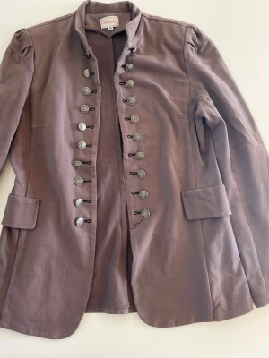 Ljusbrun/ beige kavaj med dekorativa knappar - Snygg ljusbrun/beige kavaj från chloe london med militärinspirerad design. Kavajen har dubbla rader av dekorativa silverfärgade knappar framtill, axelvaddar och två fickor med lock. Tillverkad i mjukt tyg som ger en bekväm känsla och cool look. Storleken är väldigt liten, passar mig som xs/s!!