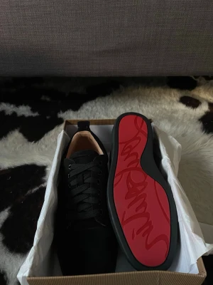 Svarta sneakers från Christian Louboutin - Snygga svarta sneakers från Christian Louboutin med klassisk röd sula och diskret logga. Skorna har en låg profil, svarta skosnören och är tillverkade i mocka. Perfekta för dig som vill ha en exklusiv och stilren look.