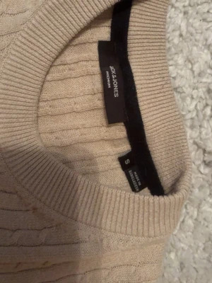 Beige stickad tröja Jack & Jones - Säljer en beige stickad tröja från Jack & Jones. Tröjan har rund halsringning, ribbade muddar och ett diskret kabelstickat mönster. Perfekt för lager på lager och enkel att matcha med olika outfits. Använd ett fåtal gånger 